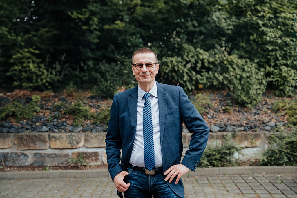 Ein Mann mit Brille, blauer Anzugjacke, gestreifter Krawatte und Jeans steht im Freien, die Hand in die Hüfte gestemmt, und lächelt - ein sympathischer Fachmann für Ihre Bedürfnisse als IT Unternehmen Dresden - vor einer grünen Kulisse und einer Steinmauer.