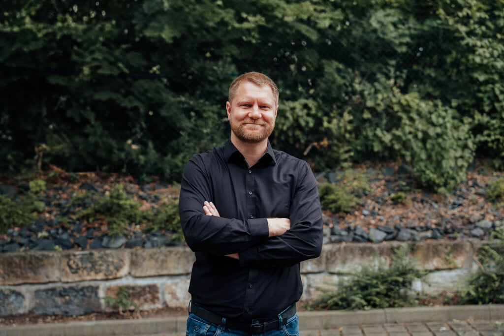 Ein Mann mit kurzen Haaren und Bart, der ein schwarzes Button-up-Hemd und blaue Jeans trägt, steht im Freien mit verschränkten Armen - er stellt einen IT-Dienstleister Dresden dar - vor grünem Laub und einer Steinmauer.