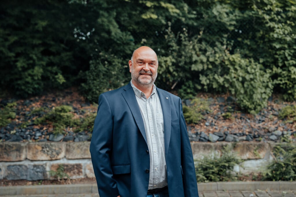 Ein Mann mit Glatze und Bart steht im Freien vor einer Grünanlage, er trägt einen dunklen Blazer über einem hellen Hemd. Er lächelt leicht und schaut in die Kamera - bereit, Ihre IT Infrastruktur als erfahrener IT Dienstleister Dresden zu unterstützen.