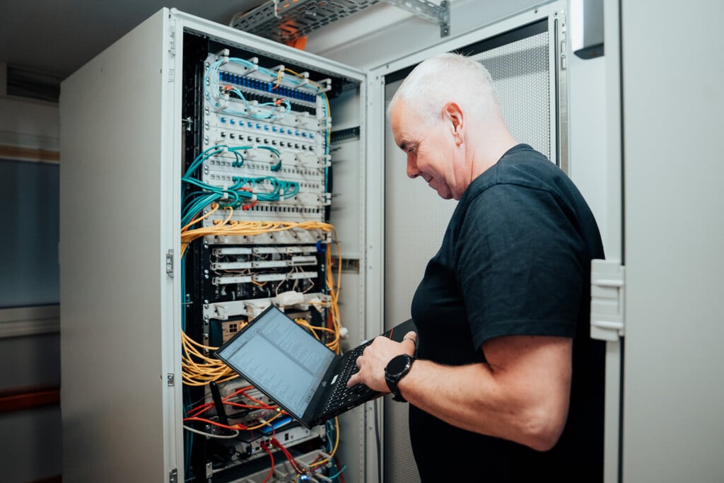 Ein Mann im schwarzen T-Shirt steht neben einem offenen Serverschrank, hält einen Laptop in der Hand und prüft Netzwerkkabel - er demonstriert die Kompetenz eines IT-Dienstleisters Dresden in einer modernen Serverschrankumgebung.