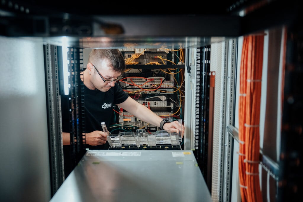 Ein Techniker, der eine Brille trägt, arbeitet an einem Server in einem Rechenzentrums-Rack, umgeben von verschiedenen Kabeln und elektronischen Komponenten. Dies zeigt die entscheidende Rolle der Netzwerktechnik bei der Wartung der IT-Infrastruktur für ein IT-Unternehmen in Dresden.