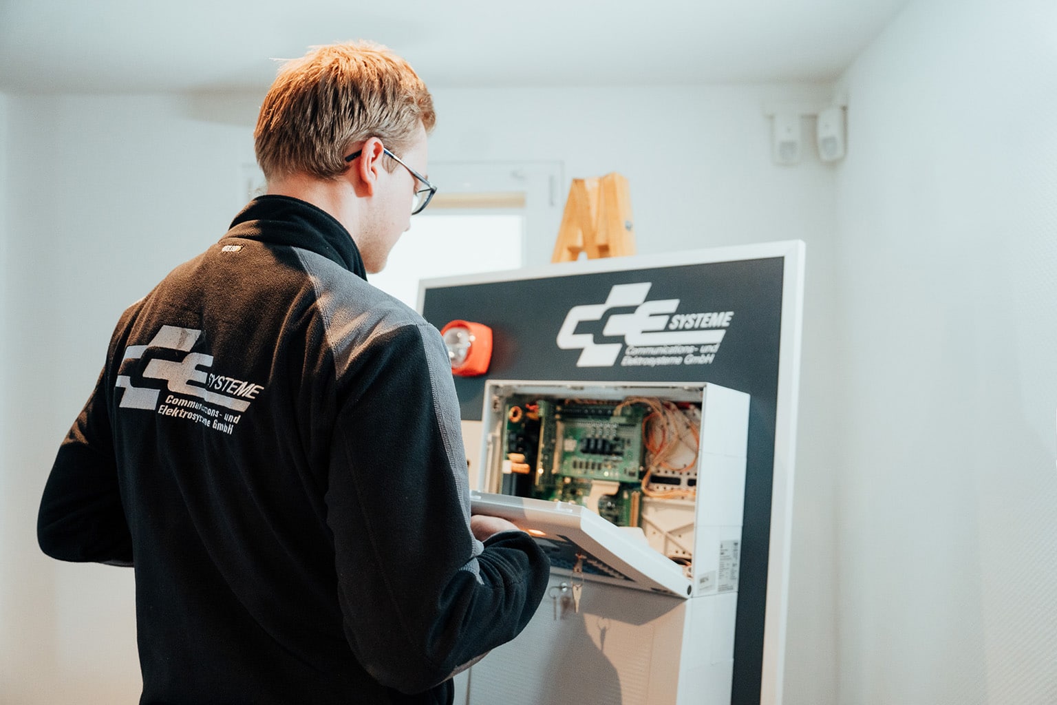 Ein Techniker mit Brille und schwarzer Jacke mit "CC SYSTEME"-Logo prüft in einem hellen, weißen Raum ein offenes Bedienfeld mit freiliegenden Schaltkreisen, um die optimale Leistung der IT-Infrastruktur und Netzwerktechnik zu gewährleisten.