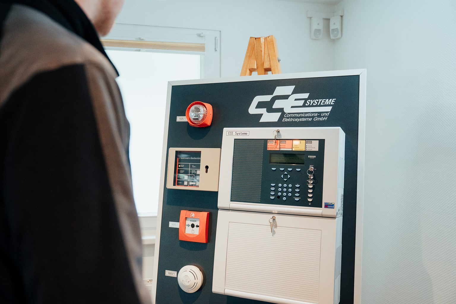 Eine Person steht vor einer Schautafel mit elektronischen Schalttafeln, Brandmeldekomponenten und dem Logo "CC Systeme", die in einem hellen Innenraum die netzwerktechnische Kompetenz eines innovativen IT-Unternehmens aus Dresden präsentiert.