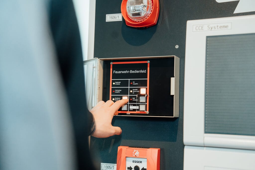 Eine Person drückt eine Taste mit der Aufschrift "Feuerwehr-Bedienfeld" auf einer Brandmeldezentrale, die Teil der fortschrittlichen IT-Netzwerklösungen ist, die von einem IT-Unternehmen in Dresden eingesetzt werden. In der Nähe sind verschiedene Tasten, Anzeigen und Sicherheitseinrichtungen zu sehen.