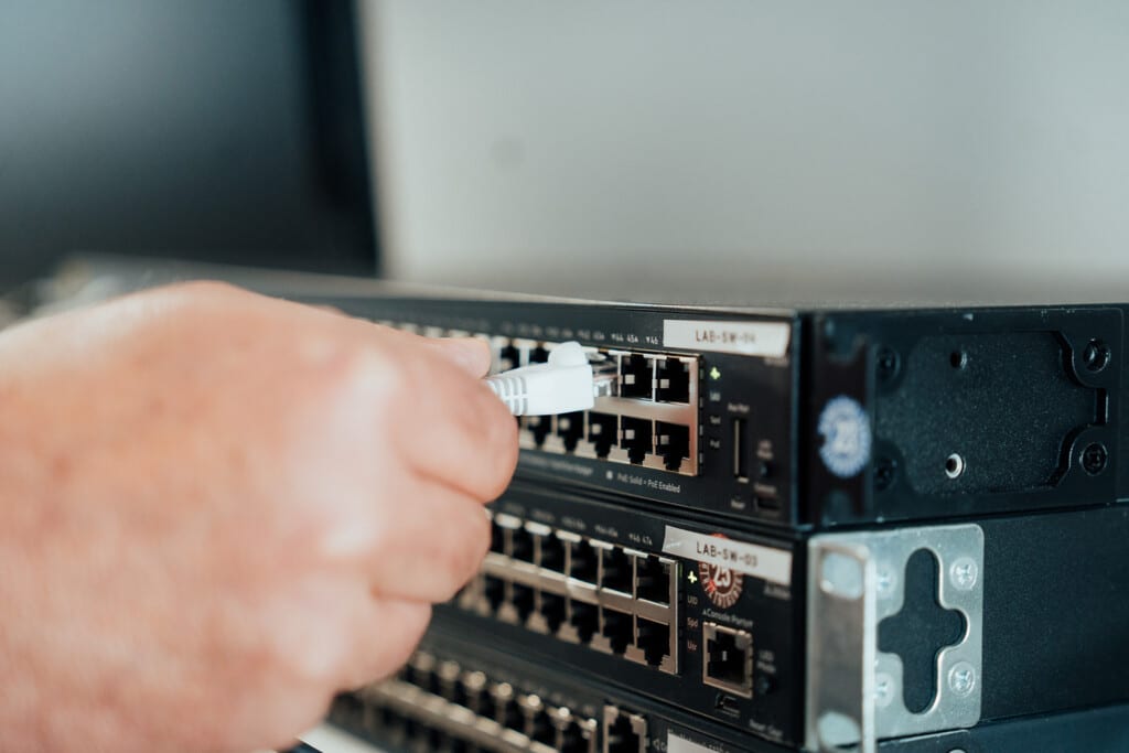 Eine Hand steckt ein Ethernet-Kabel in einen Netzwerk-Switch-Port, ein weiterer Switch befindet sich darunter in einem Rack - ein Beispiel für moderne Netzwerktechnik, die für jeden IT-Dienstleister Dresden, der eine robuste IT-Infrastruktur verwaltet, unerlässlich ist.