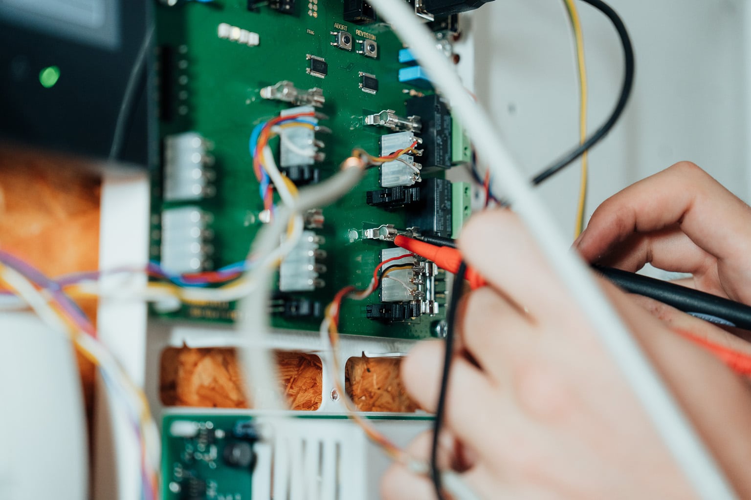 Eine Nahaufnahme von Händen, die Sonden verwenden, um eine grüne Leiterplatte mit verschiedenen Drähten und elektronischen Komponenten zu prüfen oder zu reparieren - ein Beweis für die Präzision und das Fachwissen eines IT-Dienstleisters in Dresden im Bereich Netzwerktechnik.