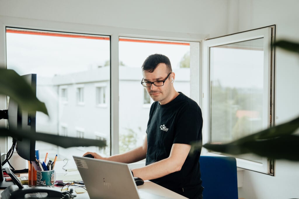 Ein Mann, der eine Brille und ein schwarzes T-Shirt trägt, arbeitet an einem Schreibtisch mit einem Laptop und Büromaterialien, neben einem großen Fenster mit offener Scheibe. Im Vordergrund sind teilweise Pflanzen zu sehen, die den modernen Arbeitsbereich eines IT-Dienstleisters in Dresden widerspiegeln.