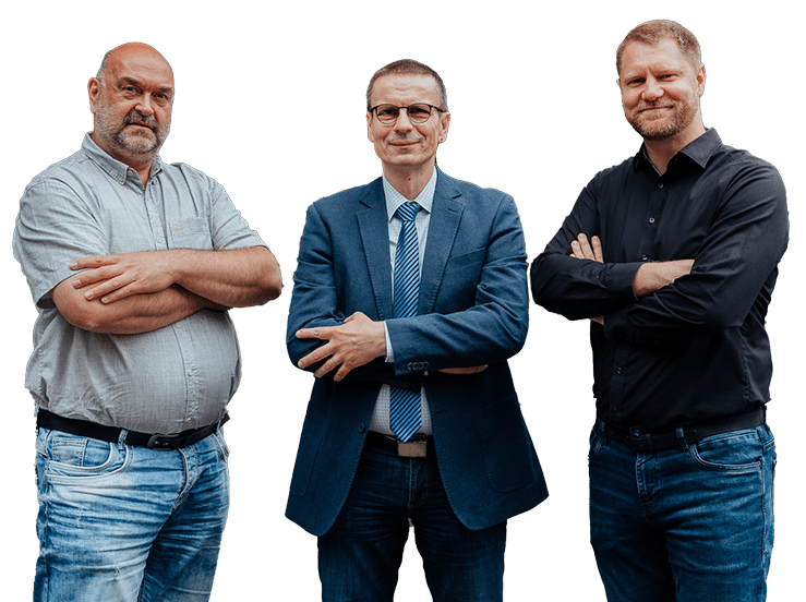 Drei Männer stehen nebeneinander vor einem weißen Hintergrund. Der Mann auf der linken Seite trägt ein graues Hemd und Jeans, der Mann in der Mitte einen Anzug mit Krawatte und der Mann auf der rechten Seite ein schwarzes Hemd und Jeans. Alle haben die Arme verschränkt.