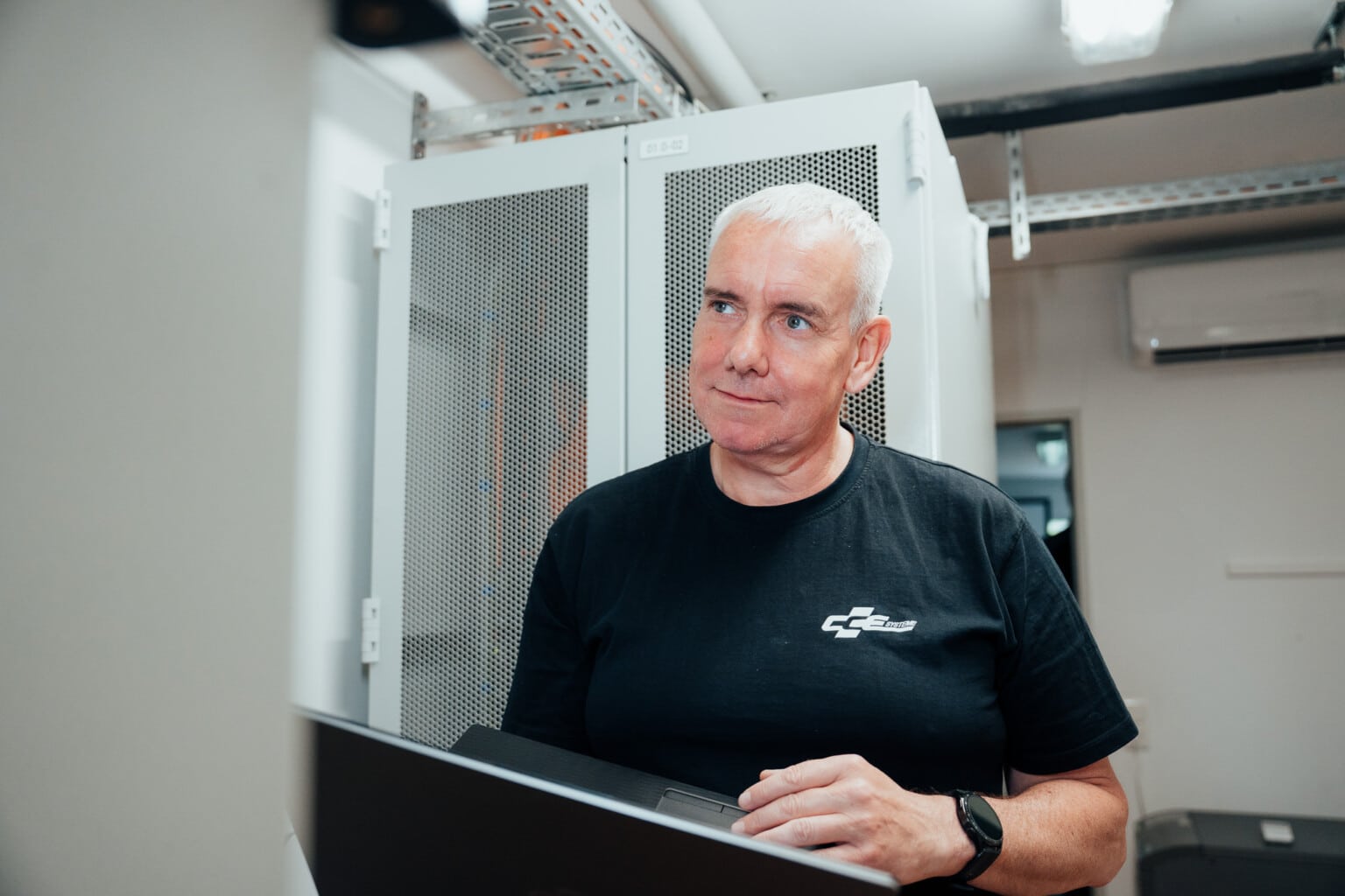 Ein Mann mit kurzen grauen Haaren und schwarzem T-Shirt steht in einem Serverraum, hält einen Laptop in der Hand und blickt leicht nach oben, umgeben von Serverschränken und moderner Netzwerktechnik, die auf eine robuste IT-Infrastruktur hinweist.